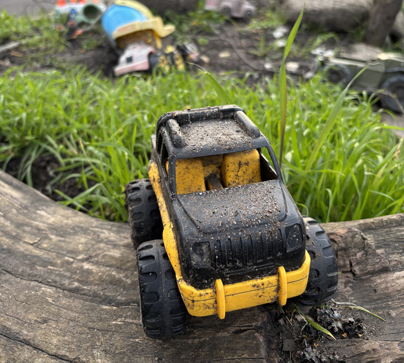 A yellow truck in the foreground; other cars and objects in a sandbox in the background.