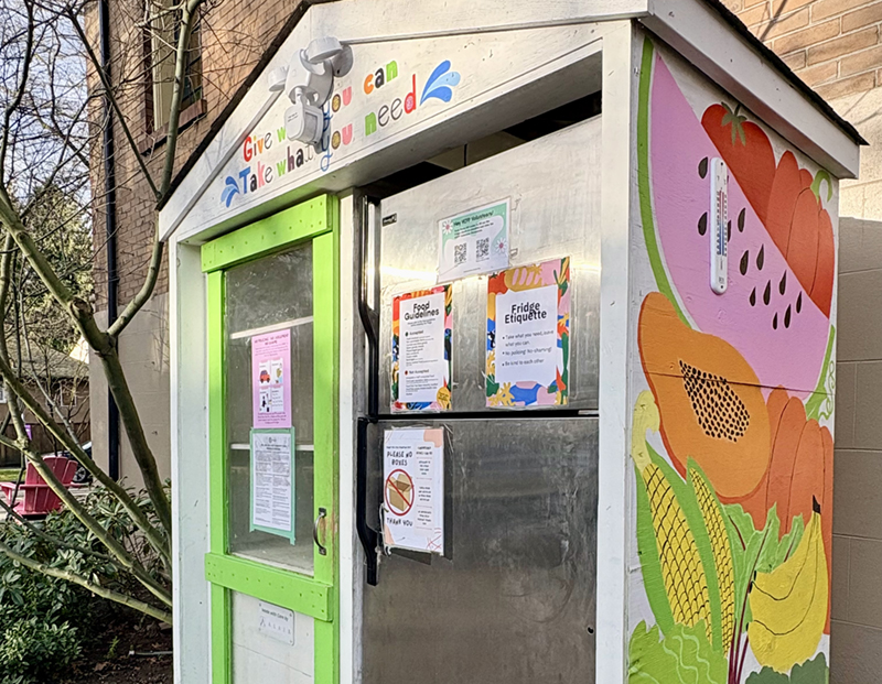 A food pantry (a shelf with green trim) and neighbourhood fridge (a stainless steel appliance) is housed in a white wooden structure. Its side to the right of the frame is painted colourfully with illustrations of fruit and vegetables.