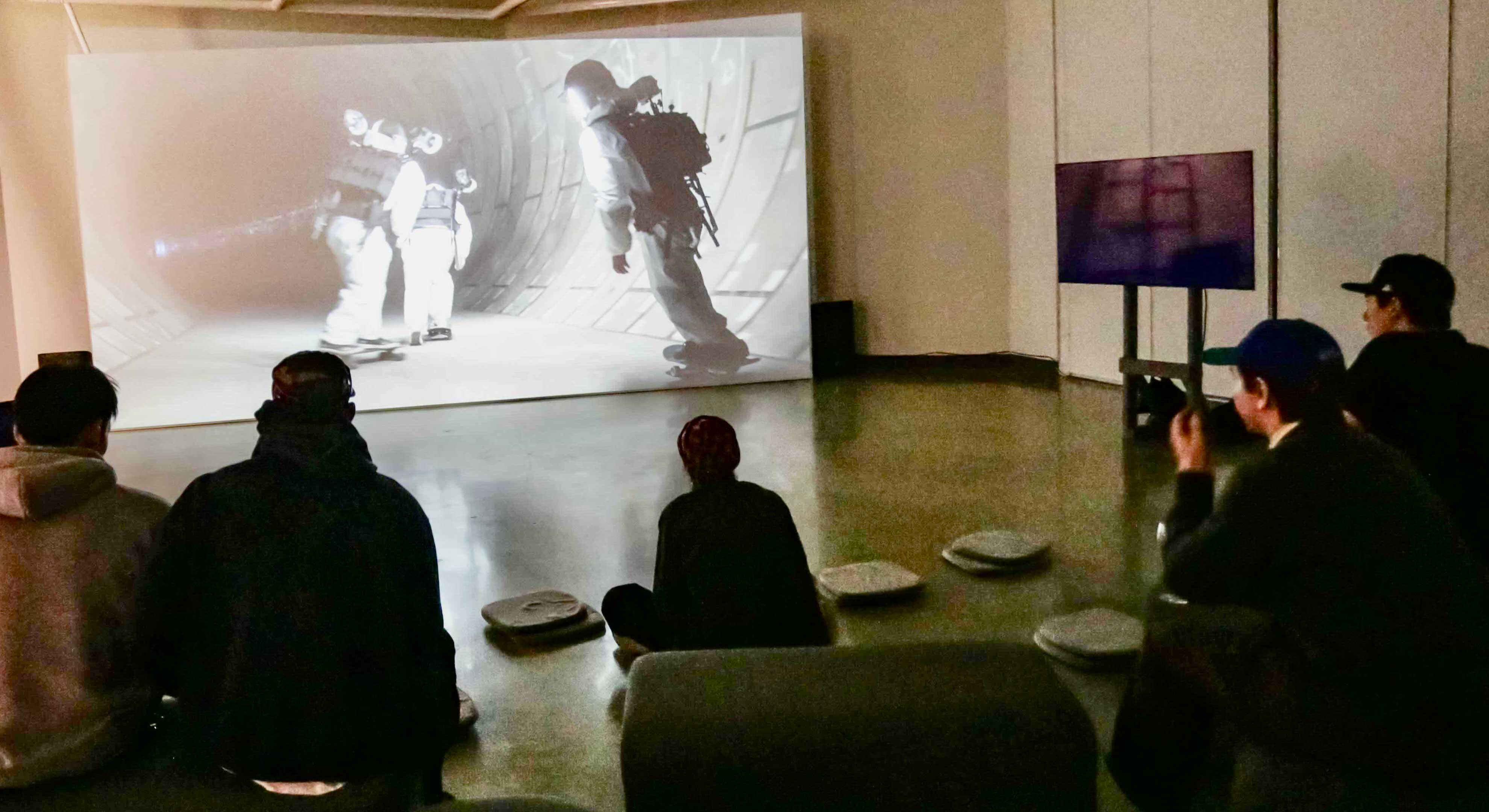 Several young people in casual streetwear and ball caps are seated on low structures in an indoor gallery space. Their backs are turned to the camera as they watch a black-and-white video installation of skateboarders in an underground tunnel on a large screen on the far wall.