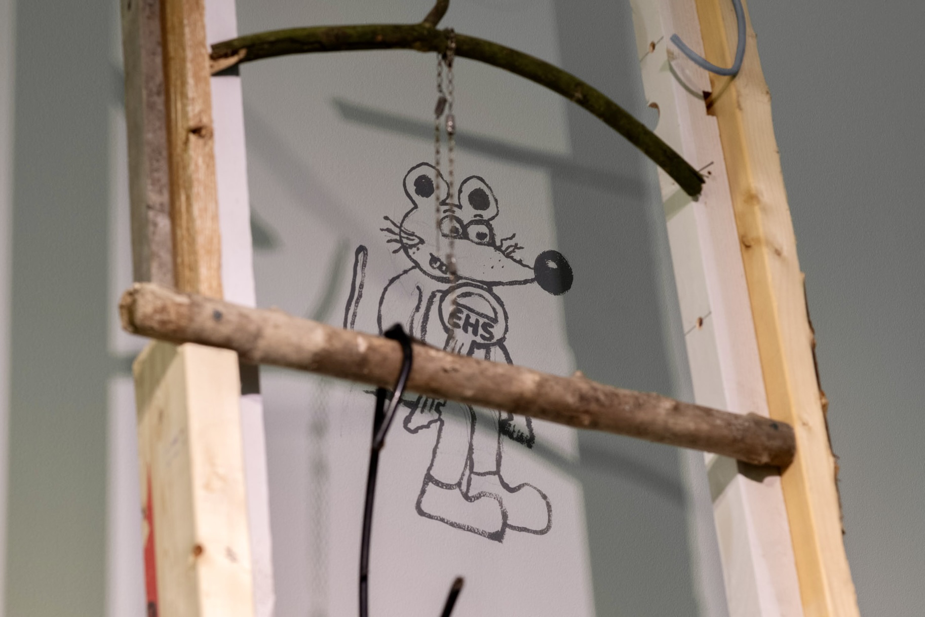 A close-up photograph of a wooden ladder in an art gallery installation that leans against a wall. The wall is inscribed with a black illustration of a cartoon rate holding a house key with the letters &ldquo;EHS&rdquo; inscribed on it.
