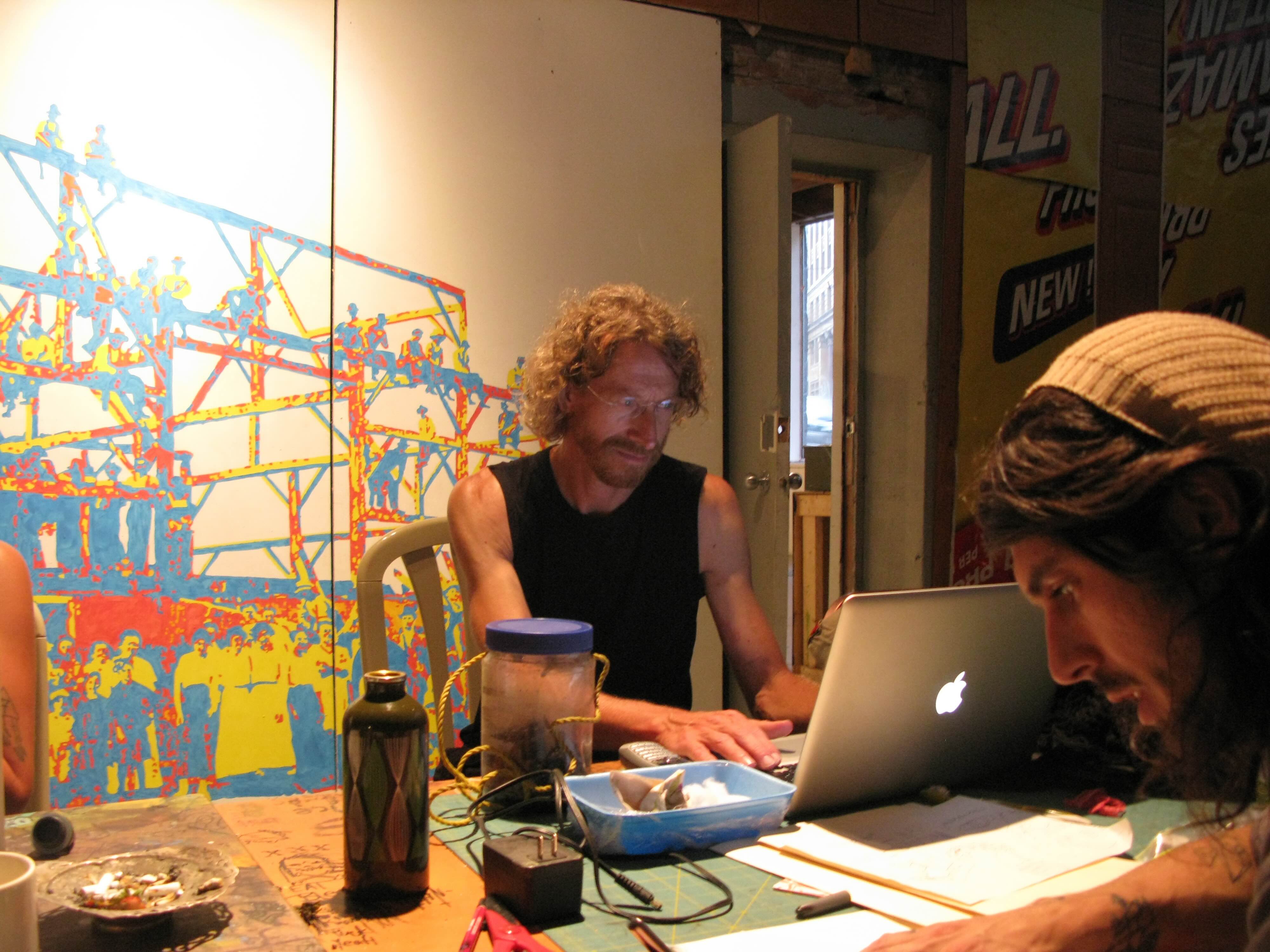 Two people work together in a dimly lit room at a table. Jim Carrico, centre, is working on a silver Apple laptop. He is wearing a black tank top and he has wavy red hair and glasses. In the foreground to the right of the frame, Jamie Biz has long wavy dark brown hair and he is wearing a beige toque.