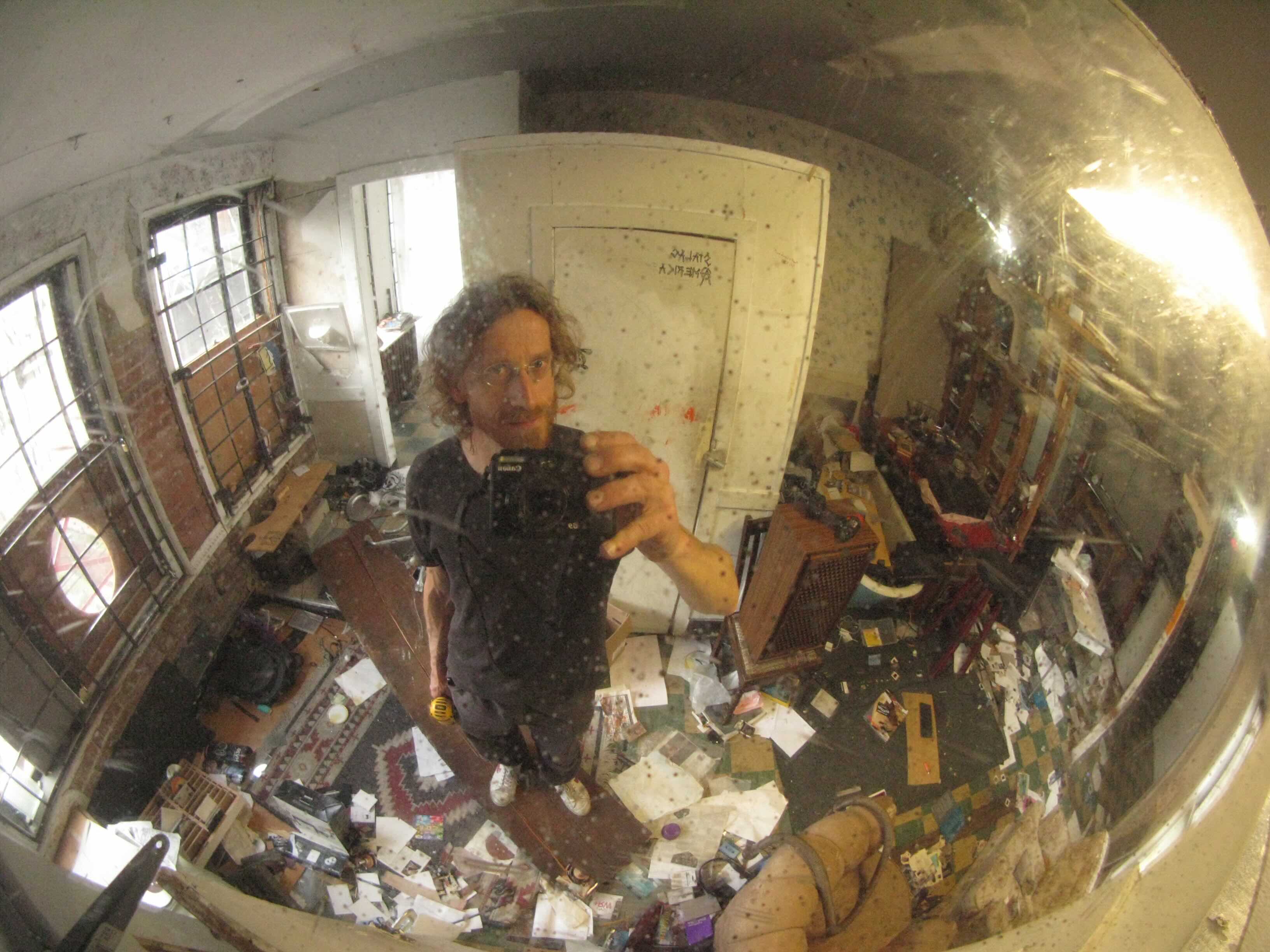 Jim Carrico has wavy red hair and glasses. He is standing holding a camera towards a convex mirror which depicts him standing in a small room littered with refuse. 