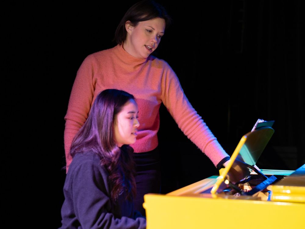 Andrea Unrau stands behind Angelaica Morse seated at a yellow piano on a darkened stage. Unrau has long brown hair tied back, and she is wearing a pink sweater. Morse has long dark hair and she is wearing a black top.