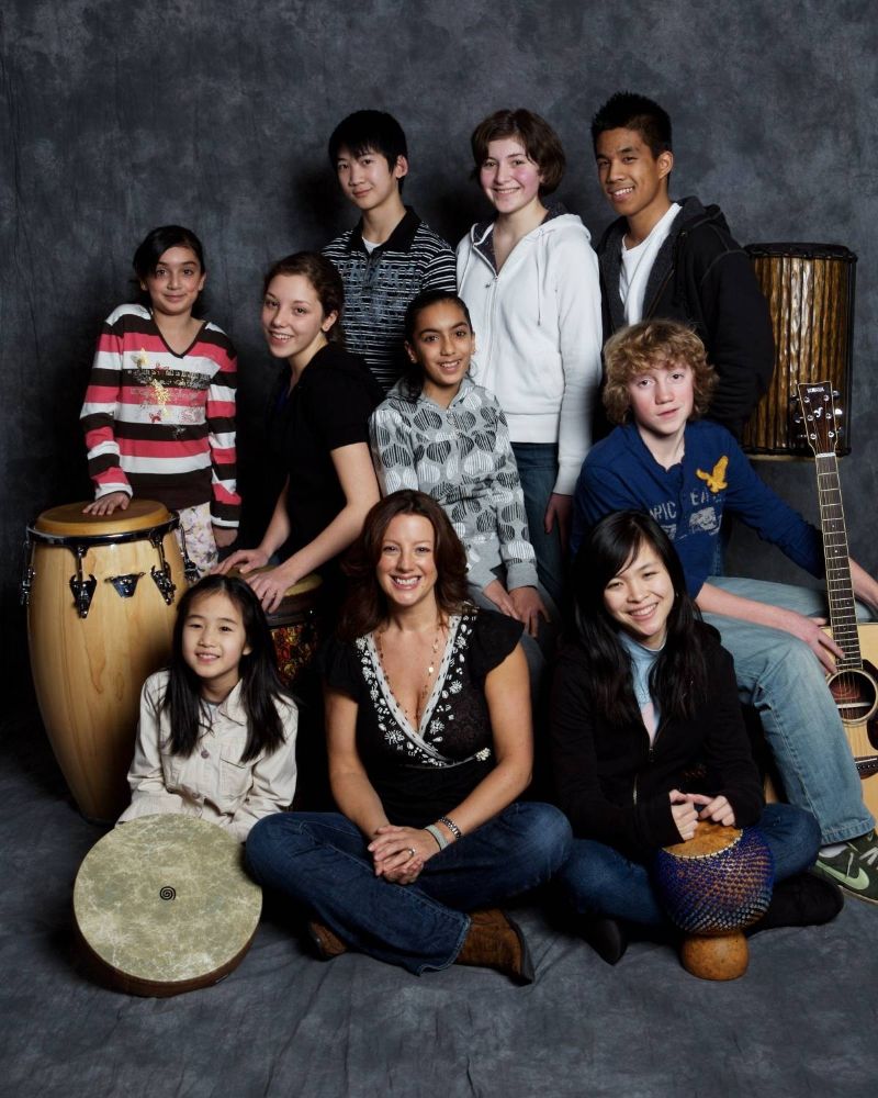 Sarah McLachlan, seated at centre, has brown shoulder-length hair. She is sitting on the floor, cross-legged, wearing jeans and a black top with white floral trim. She is smiling, and a group of students in middle and high school wearing casual streetwear are seated around her, some holding percussion or string instruments. They are posing against a grey studio background.