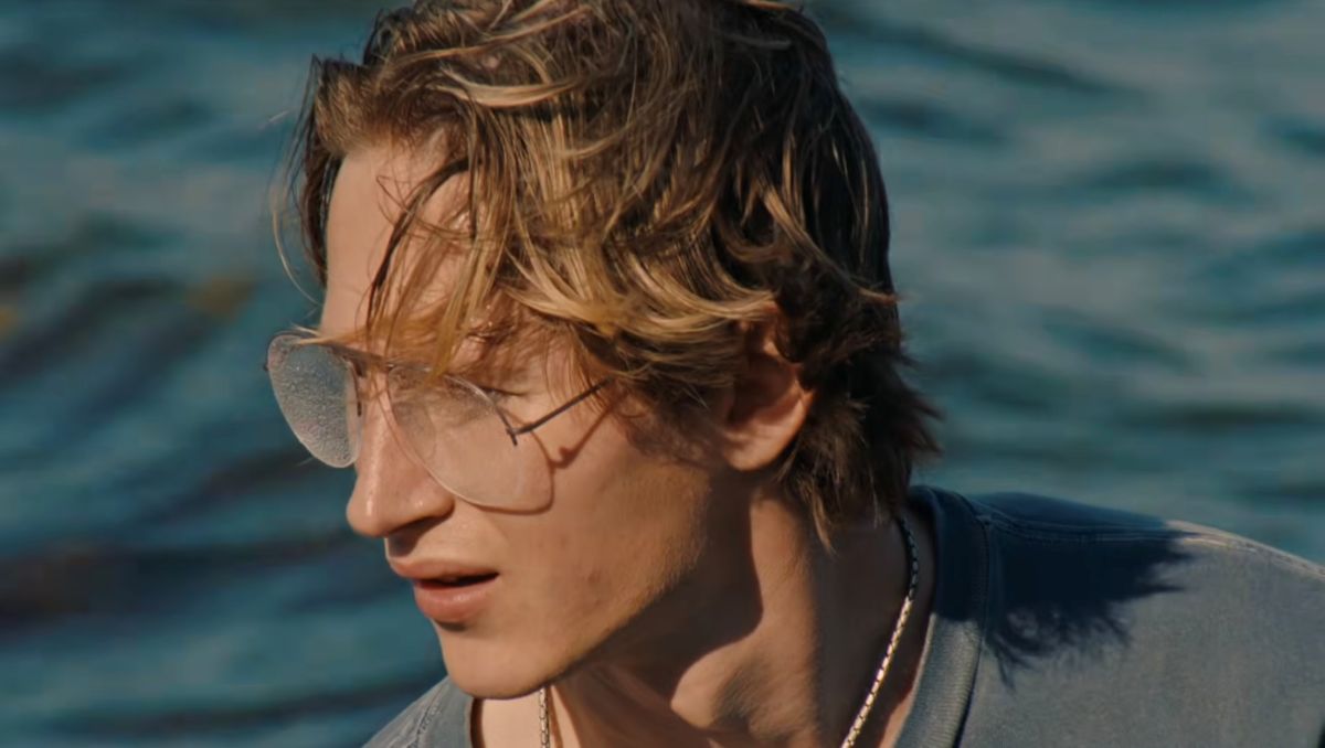 A young man with straight short blond hair and glasses looks down towards the left of the frame. He is wearing a light blue T-shirt against a blue background.