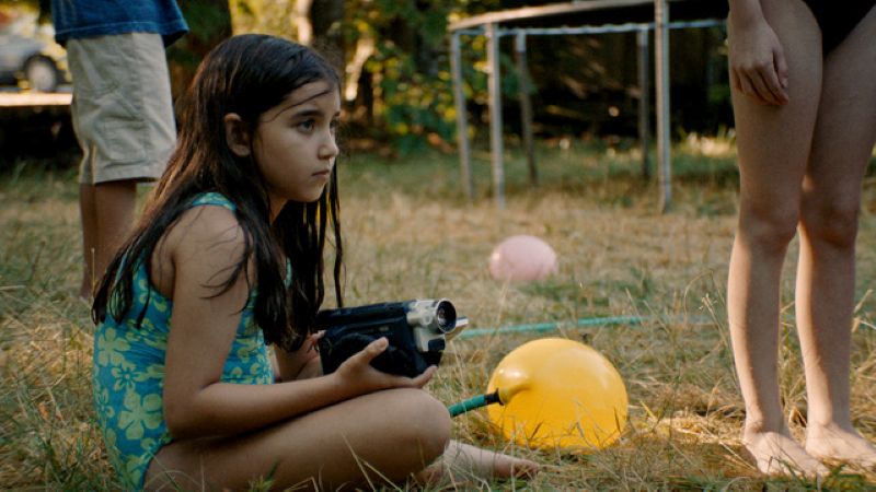 A young girl with long brown hair is wearing a blue and light green bathing suit with a floral pattern. She is sitting with her legs crossed on a lawn. She is holding a camcorder in her lap. It points to the right of the frame. The legs of other young people are around her.