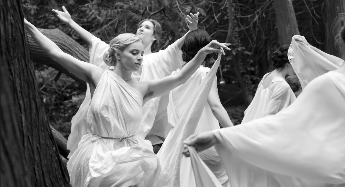A black-and-white photo depicts group of young women wearing tunics fashioned from white bedsheets. They are dancing around a circle together outdoors.