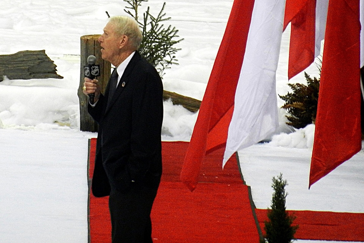 An older white man with light-coloured hair wears a suit. He is outdoors, holding a microphone, standing on a red carpet.