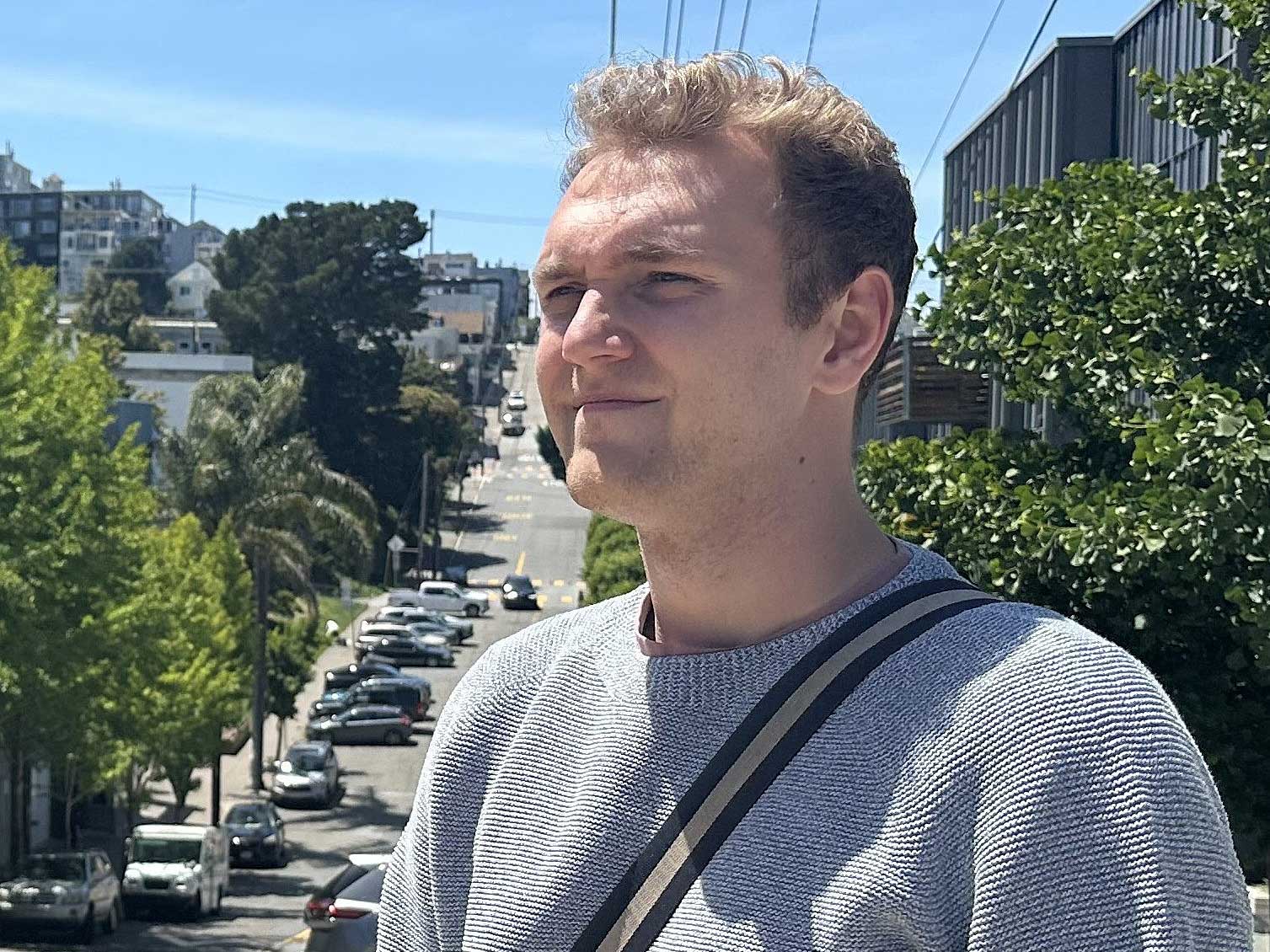 Liam Hebert has short curly blond hair. He is looking towards the left of the frame, into the middle distance. He is wearing a light grey sweater and a satchel over his chest. Behind him is a hilly city street lined with green trees on a bright day.