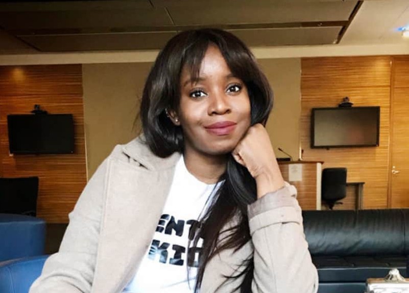 Takara Small is seated in an indoor space. Her chin is resting in one hand. She has long dark hair and is wearing a beige jacket over a white T-shirt with black lettering. Behind her are black sofas and wood-paneled walls fitted with black screens.