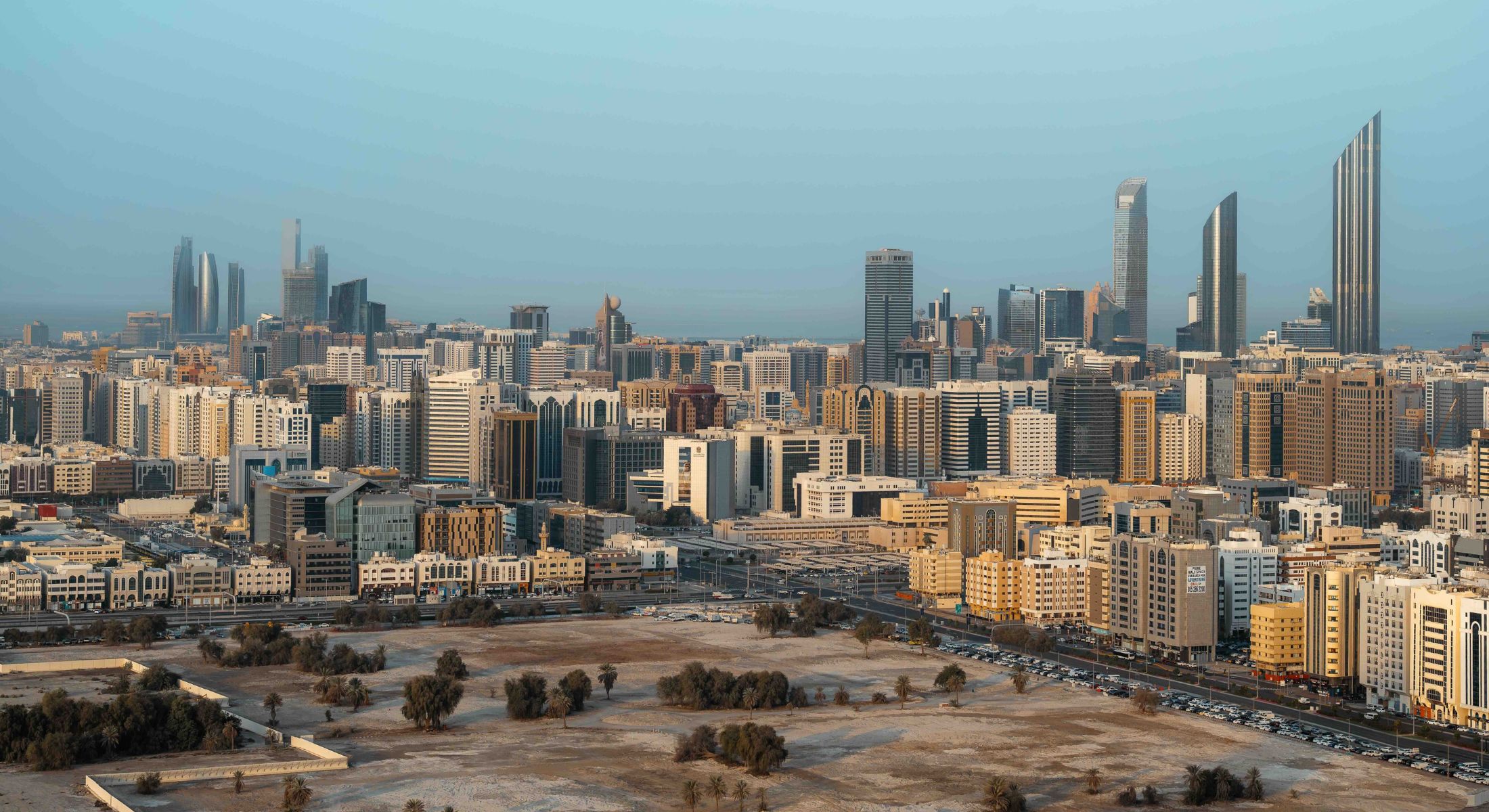 A panoramic photograph features an expansive urban landscape against a blue sky. In the foreground is a dry greenspace dotted with short trees. Around it is a dense assortment of low-rise and high-rise towers, with the tallest towers in the background.