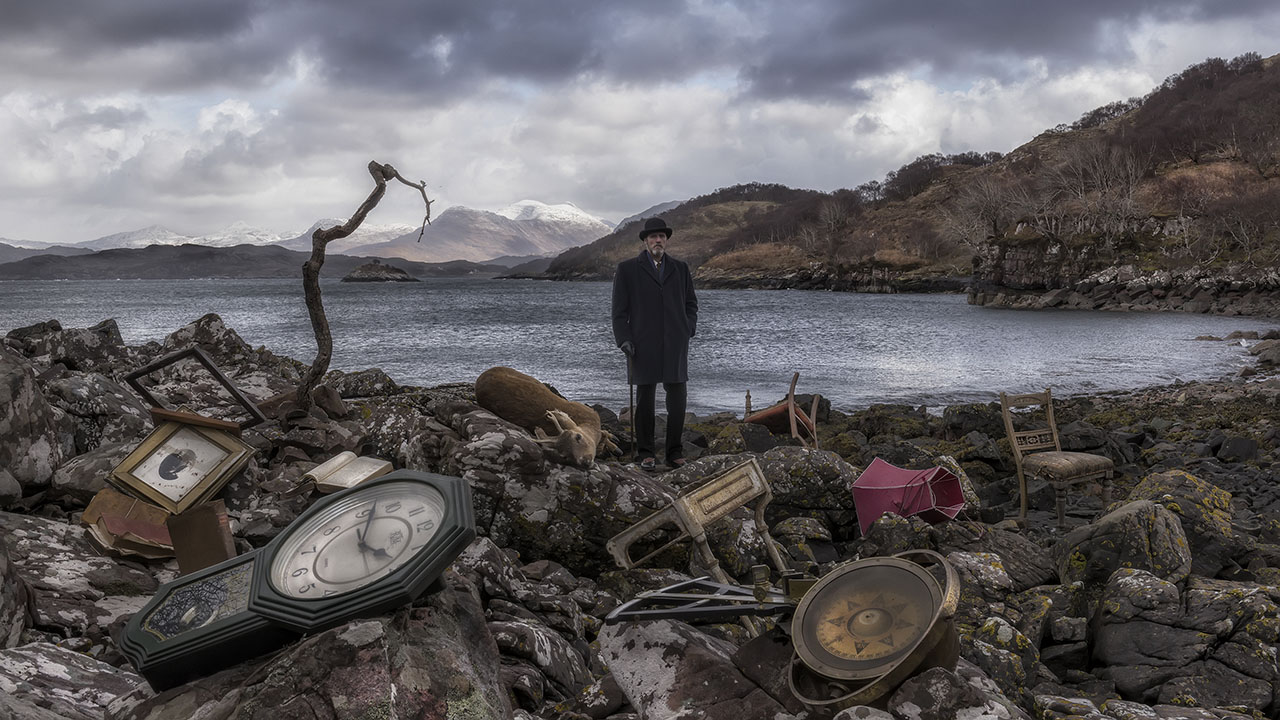 A film still features a bearded older man in a black bowler hat and dark suit standing on a rocky shoreline with the ocean, mountains and a grey sky behind him. The beach rocks around him are littered with debris including a deer carcass, a grandfather clock, old furniture and a book.