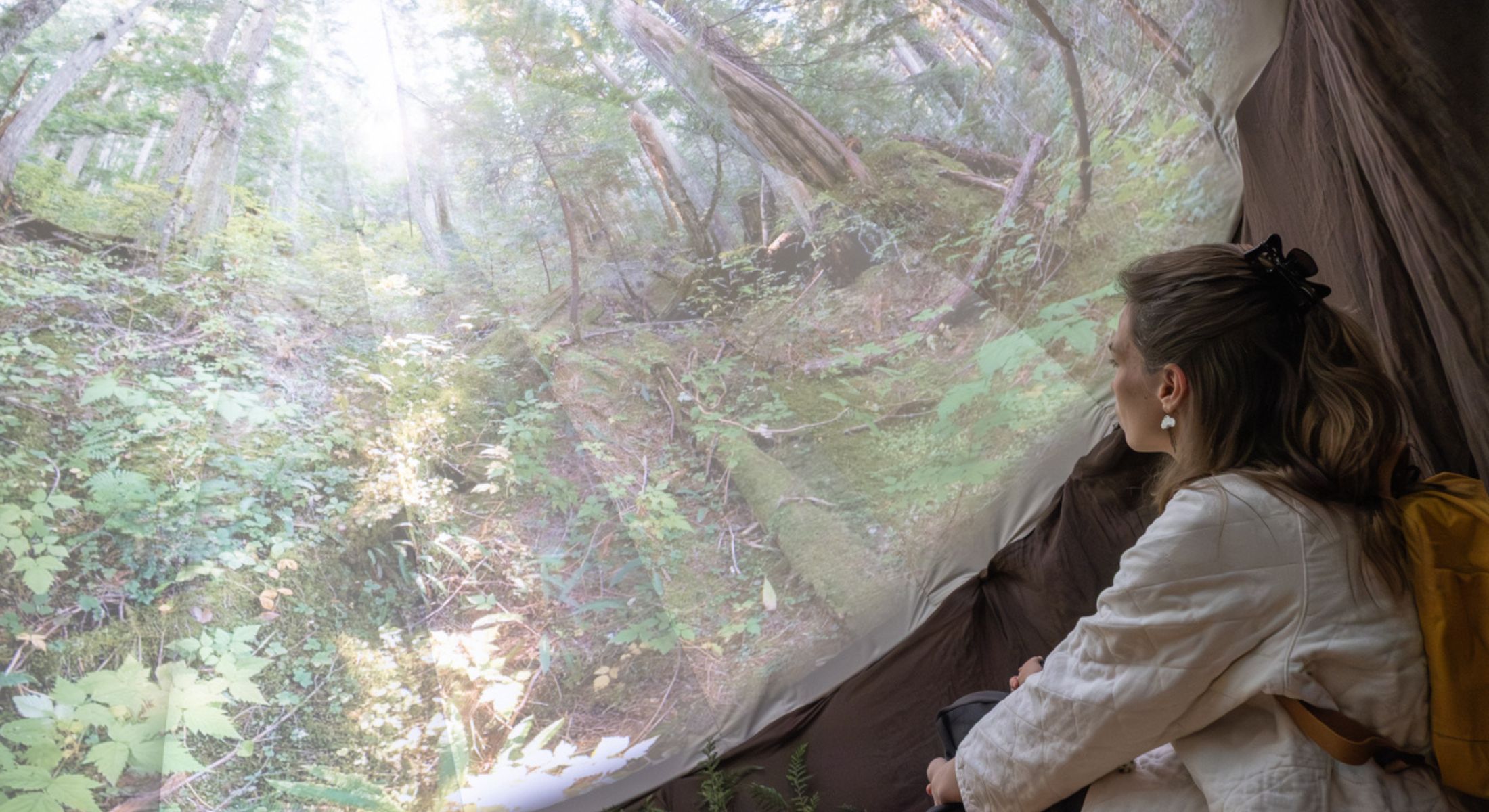 A woman is crouching to the right of the frame, and she is viewed from the side and back. She has long wavy blond hair pulled back in a clip and she is wearing a white quilted jacket. She is looking at a projection of a photograph of a forest on a round radial fabric in an indoor gallery space.