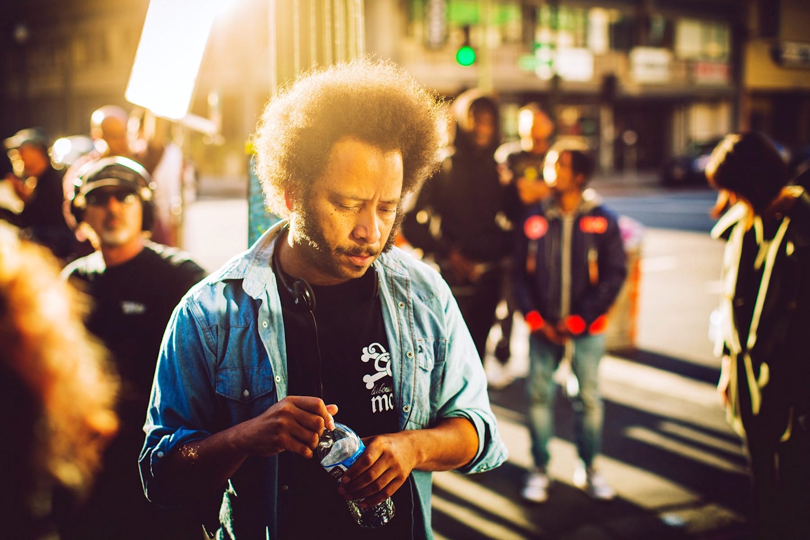 Boots Riley has curly black hair in an afro, a moustache and long sideburns. He is wearing a light denim button-down over a black T-shirt. He is looking down, holding a water bottle while standing outdoors on a film set on a city street. Behind him are lights and a film crew.