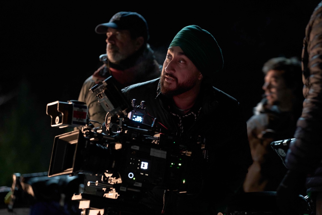 Jasmeet Raina stands behind a camera on a television set at night, looking up. He wears a dark green turban and a black jacket. Several crew members are behind him.