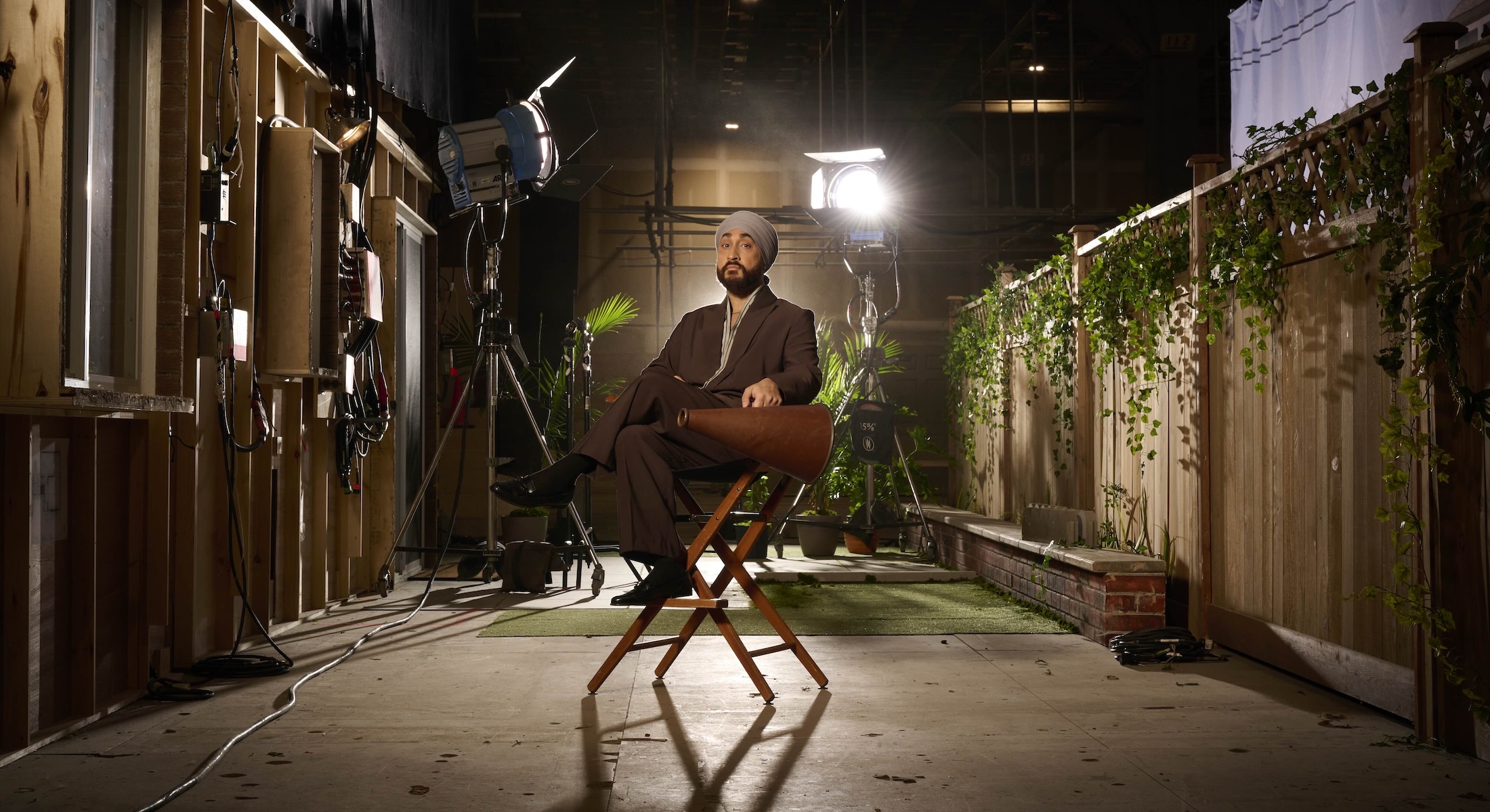 Jasmeet Raina is wearing a dark brown suit over a grey striped button-down shirt. He is sitting in a director&rsquo;s chair on a soundstage. He is holding a brown director&rsquo;s megaphone and looking at the camera, eyebrows raised. He has a dark beard and he is wearing a light grey turban. Behind him are bright studio lights and part of a TV set featuring a wooden fence hung with vines, reminiscent of what one would find in a residential backyard.