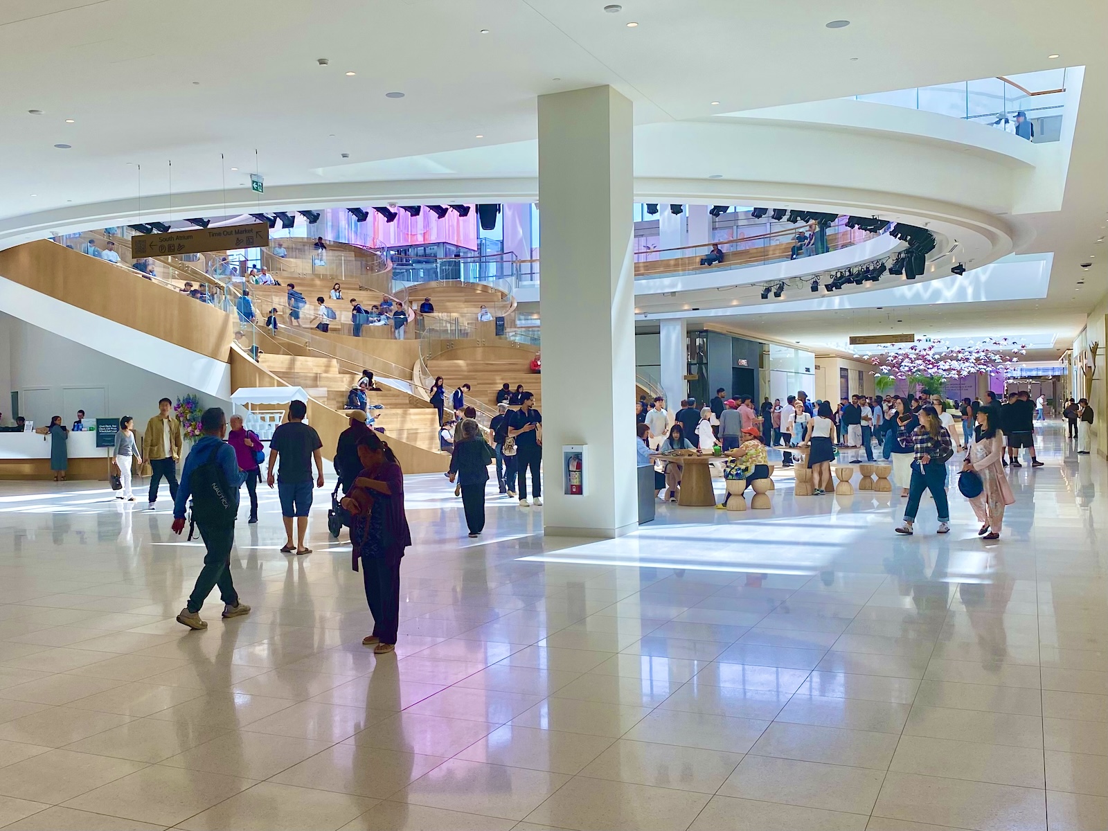People of all ages walk through and linger in an indoor concourse of a shopping mall. The walls, floors and ceilings are white. In the background is a wooden terraced seating area.