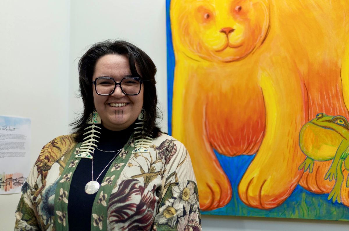 Shay Paul poses in front of a large painting of a yellow bear and toad. She has short dark wavy hair and glasses, fish skeleton earrings and a floral top over a black turtleneck. She&rsquo;s smiling.