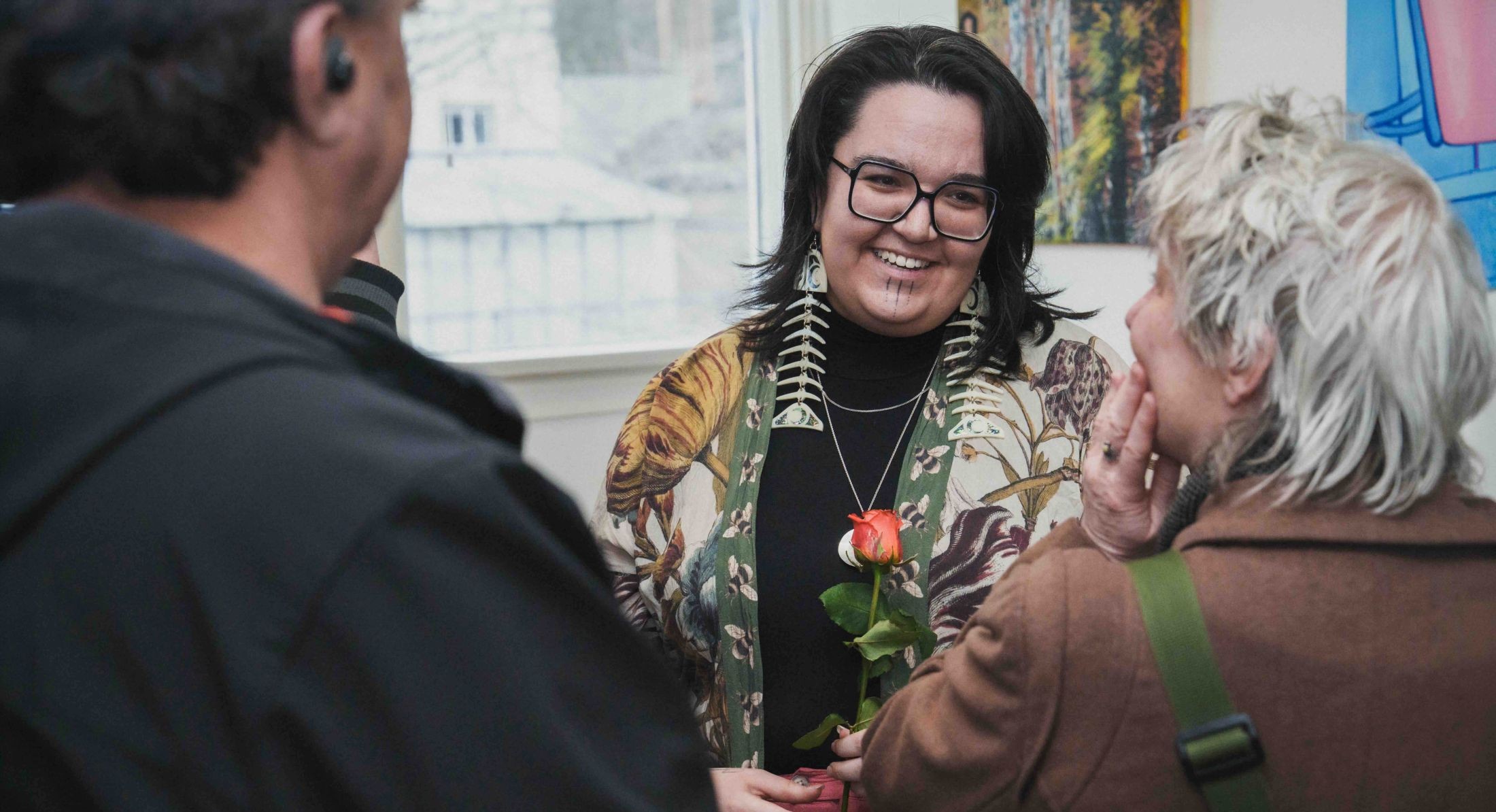 Shay Paul stands in a crowded gallery space where people are standing in conversation. She has wavy dark shoulder-length hair. She is wearing glasses and large earrings in the shape of fish skeletons. She is looking towards a person with their back turned to the camera and smiling. She is wearing a floral top over a black turtleneck and holding a rose.