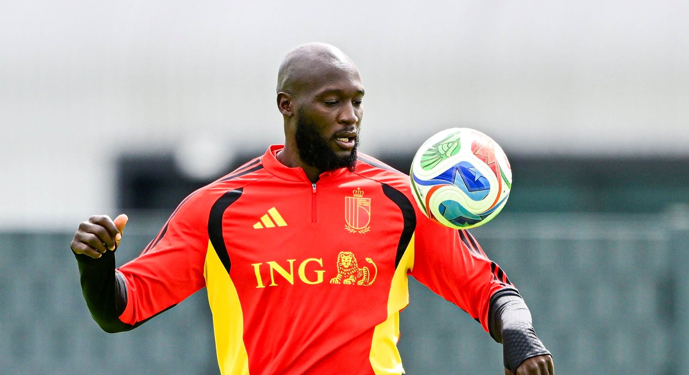 Romelu Lukaku wears a red soccer track jacket with yellow and black trim. He is looking down and right towards a colourful soccer ball in the air in front of him.