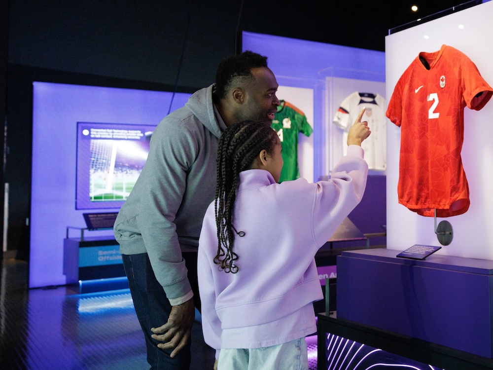A child with long dark hair in cornrows has their back turned to the camera. They are pointing to a red soccer jersey in a museum display case. An adult man leans near them nearby, smiling.