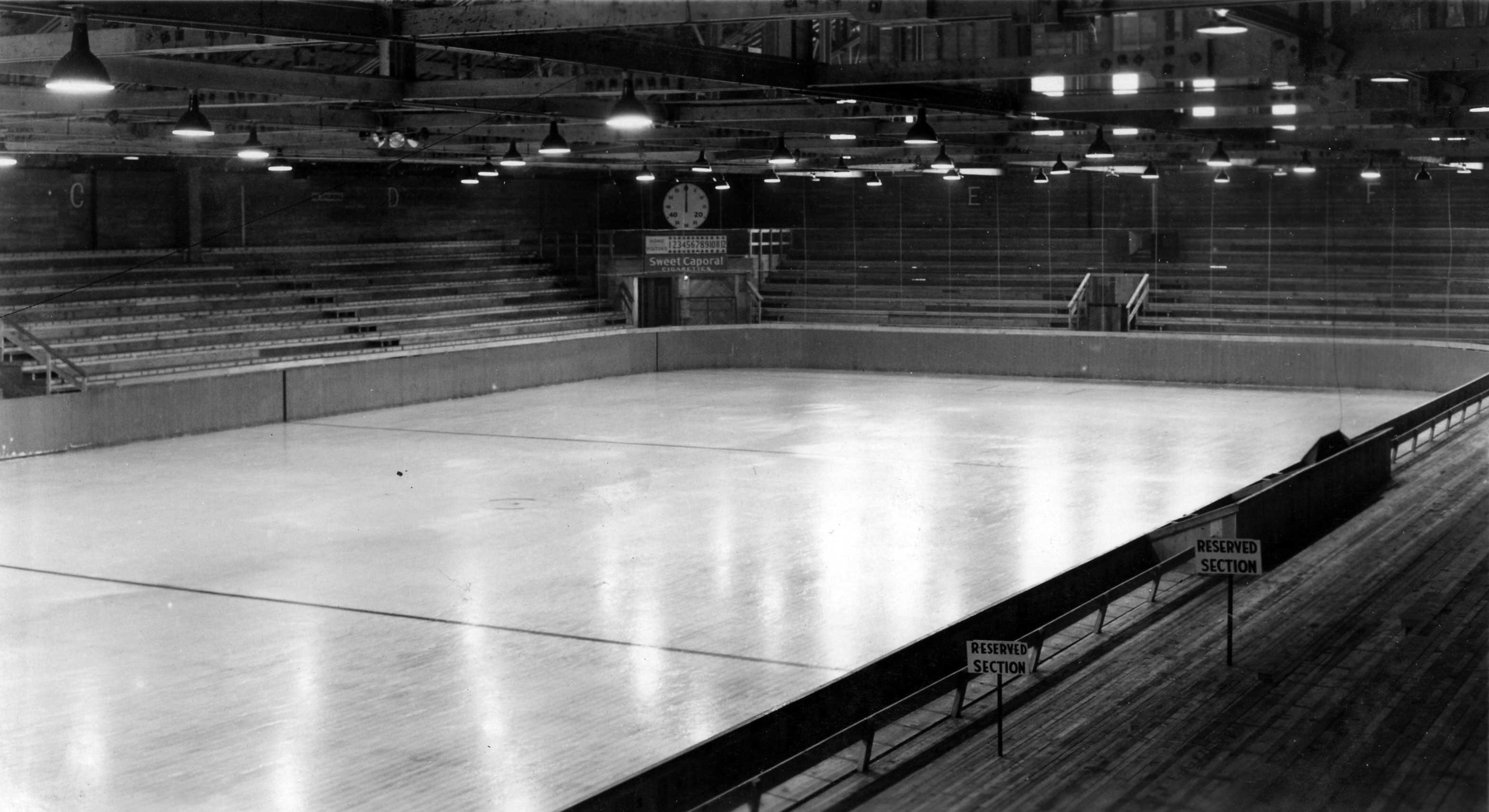 A black-and-white archival photograph depicts a hockey rink with a clock and scoreboard in the far corner, and a wooden balcony in the foreground to the lower right of the frame featuring a sign that reads &ldquo;reserved section.&rdquo;