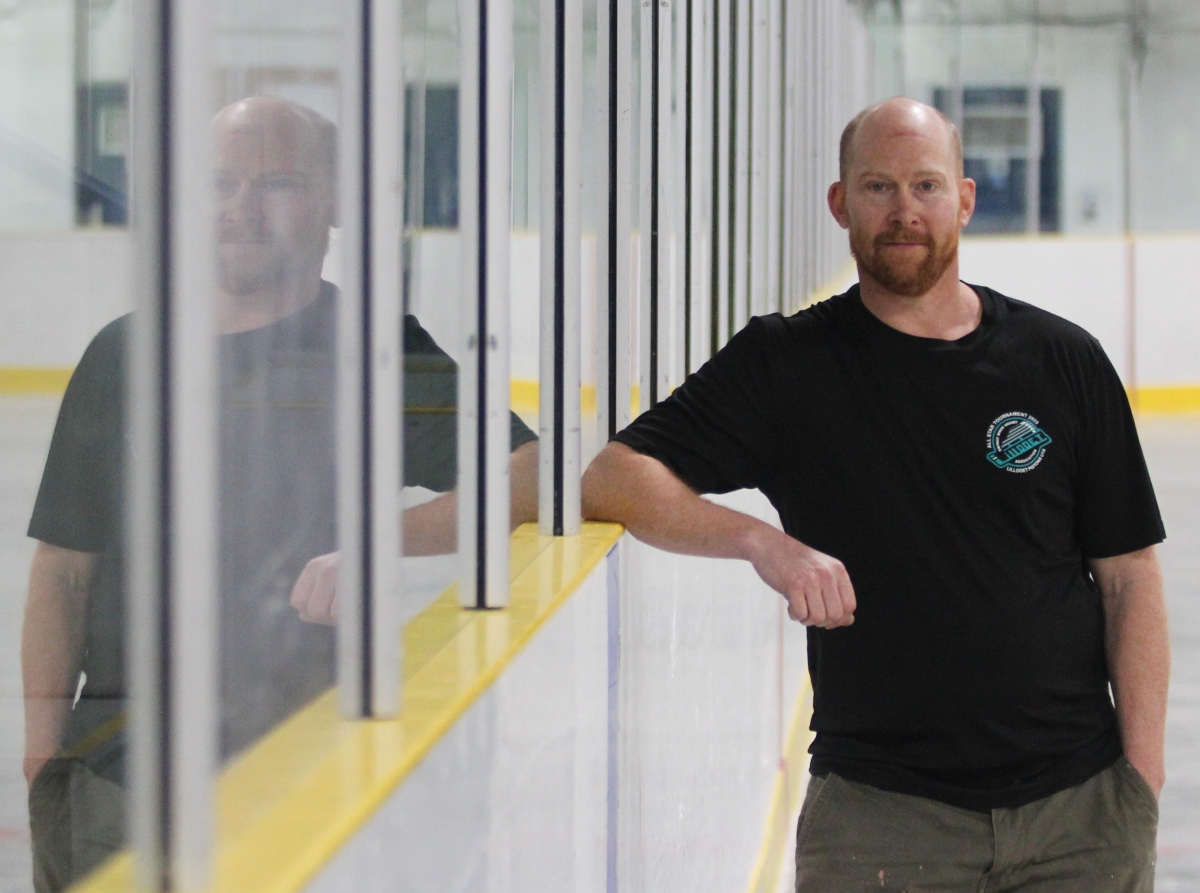 Gavin Smith stands to the right of the frame. He is wearing a black T-shirt and rets his elbow against the yellow ledge atop the boards of a hockey rink in Lillooet.