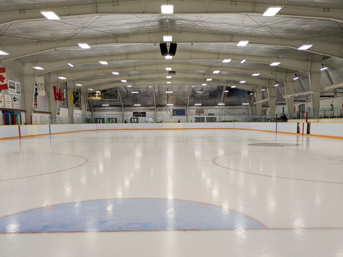 An interior photograph of the hockey rink in the Logan Lake Recreation Centre. There are fluorescent lights on the ceiling and a simple set of flags line the walls to the left of the frame.