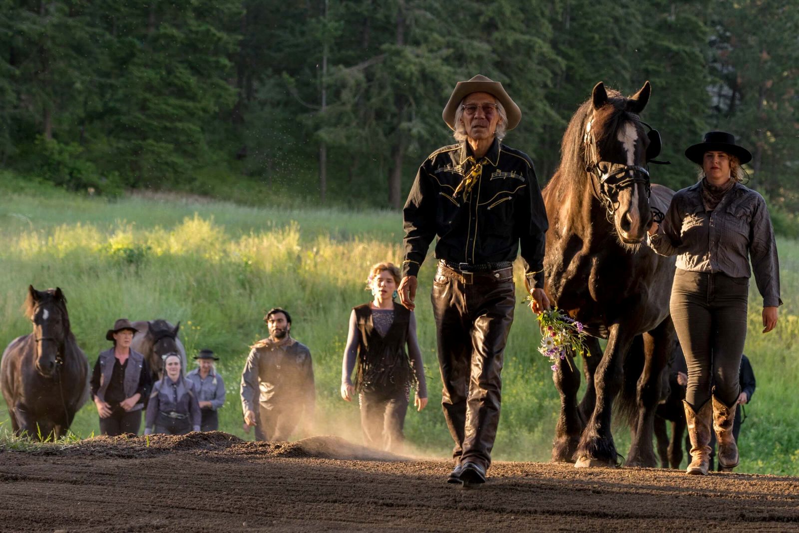 Actors in dark vests, shirts, hats and cowboy boots walk towards the camera on a dirt walkway. They accompany several dark horses as part of an outdoor theatre production. Behind them is tall grass in golden light and a stand of trees.