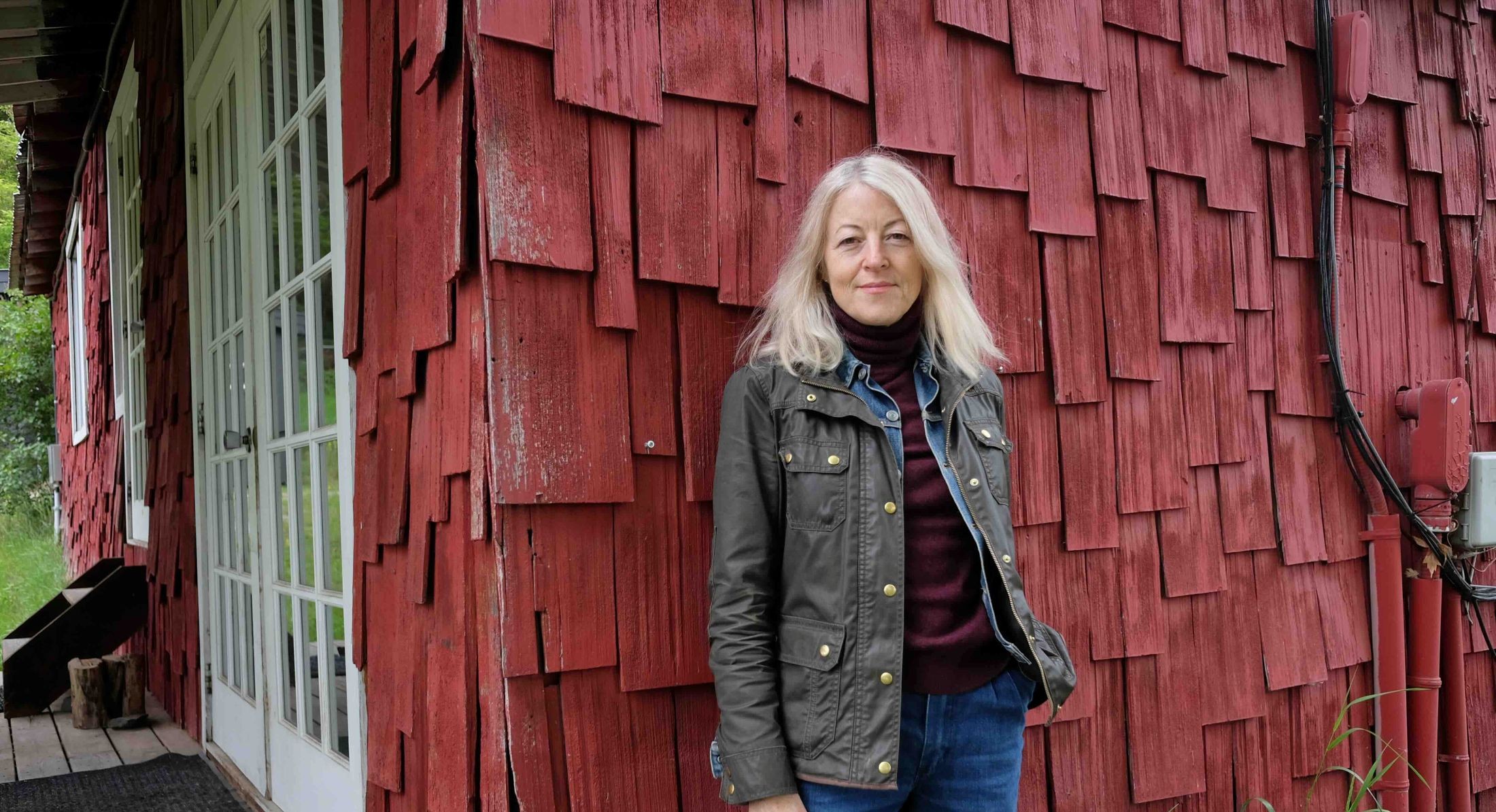 Estelle Shook stands outdoors against a shingled red wall. She wears a dark brown jacket over a denim shirt, blue jeans and a purple sweater. She has light, shoulder-length hair and she is looking directly towards the camera.