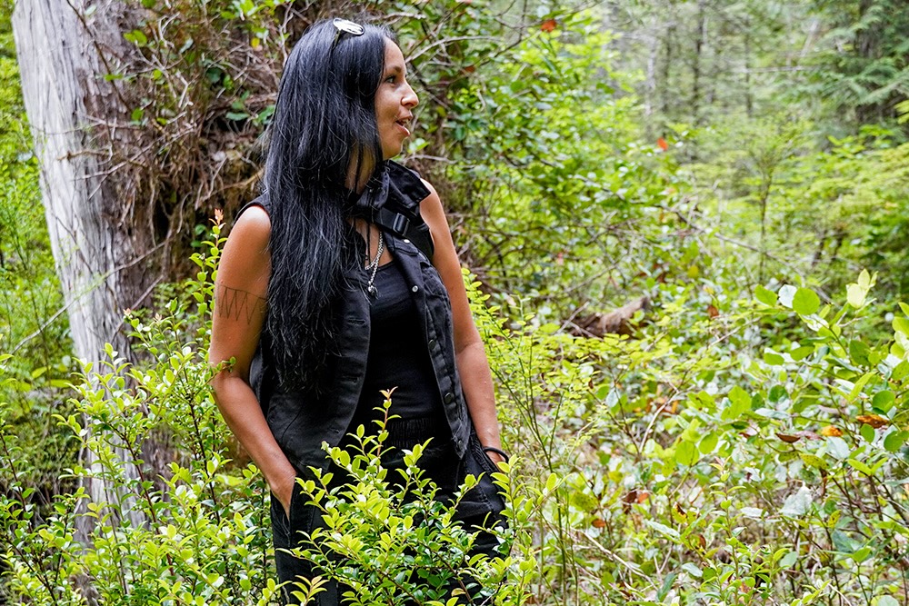 A woman with medium-light skin tone and long black hair stands among greenery in a forest.