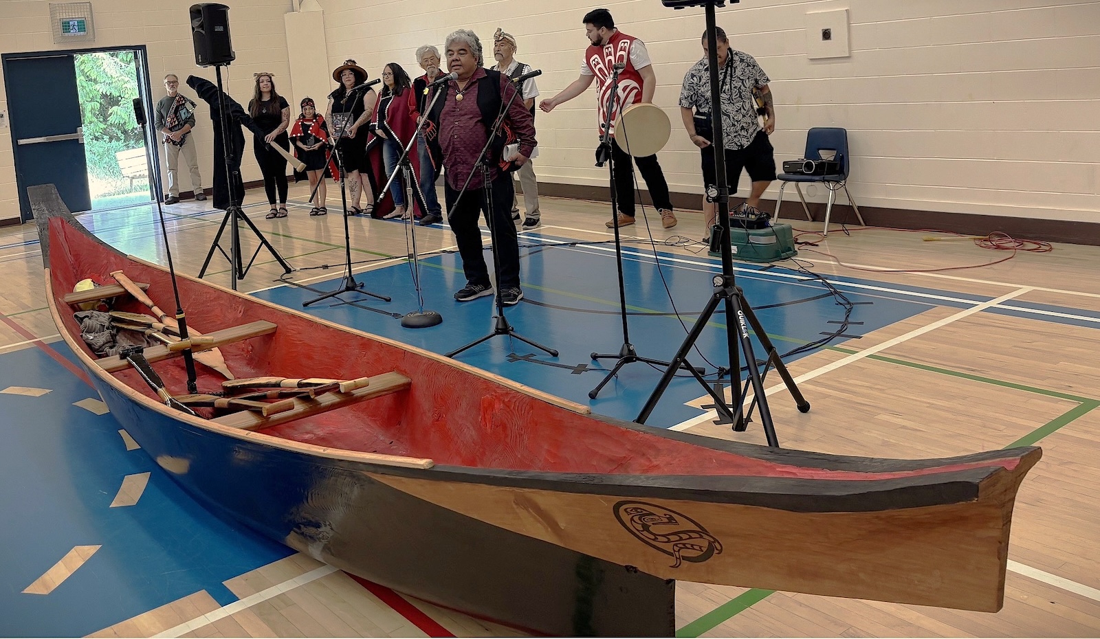 An Indigenous many in a black vest speaks into a microphone as several people watch behind him. In front of him is a dugout canoe.