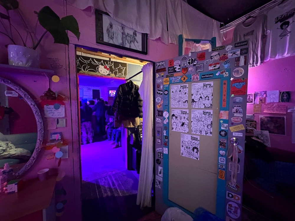 The dim interior of a DIY arts venue space whose walls are covered with stickers, comics, textiles and illustrations. A door opens to a blacklit space. The room in the foreground is dimly lit with purple hues.