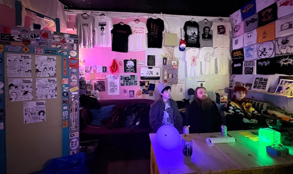 Three young adults sit together on a sofa in the lounge of a DIY venue space. Its walls are lined with T-shirts, stickers and other artwork. The space is lit dimly with purple lamps.