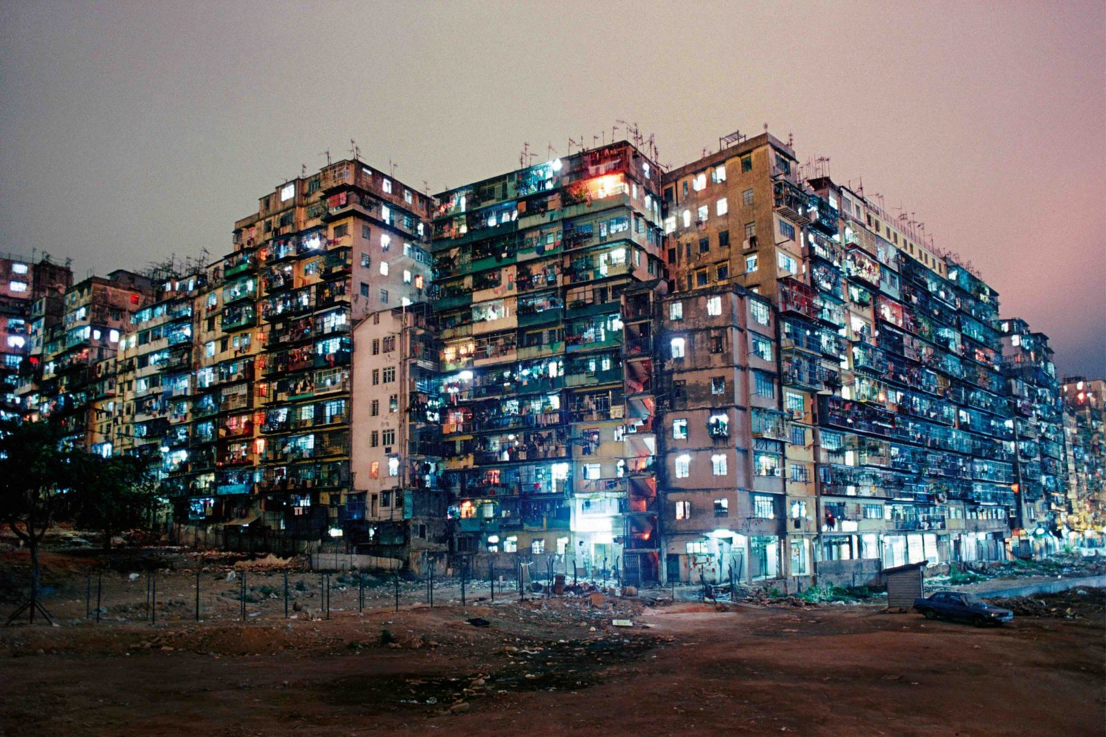 A panorama photograph of Hong Kong&rsquo;s Kowloon Walled City features a large highrise residential development lit from within at night.