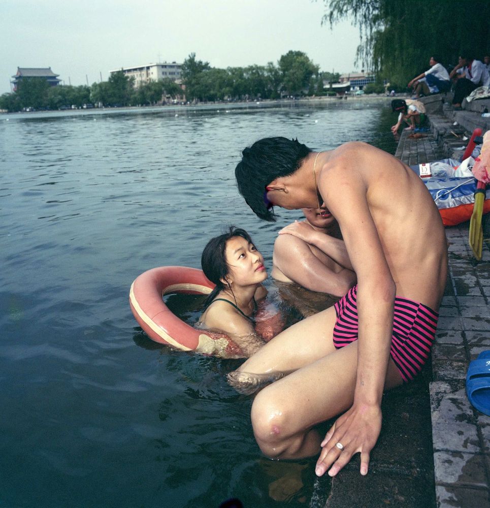 Young people gather on the concrete ledge leading to the water in Beijing. A young woman swimming in a life raft looks up to a young man seated on the ledge in a pink and black striped swimsuit.