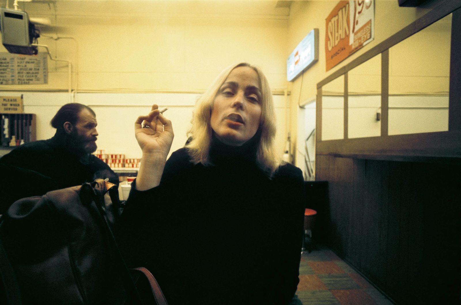 A woman with blonde hair and a black sweater holds a cigarette in one hand, exhaling. She is seated in an indoor space with light yellow walls. Behind her is a man with a beard looking towards the right of the frame.