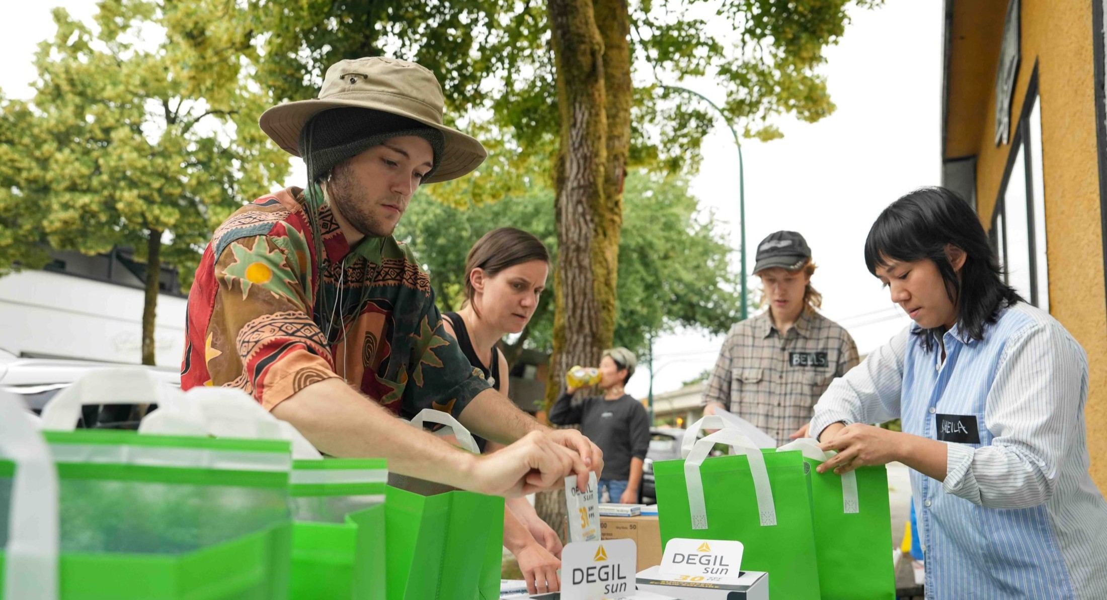A small group of people packs items into bright green bags with white handles at a long table outdoors on a sidewalk. They are wearing casual summer clothing on a clear, overcast day.