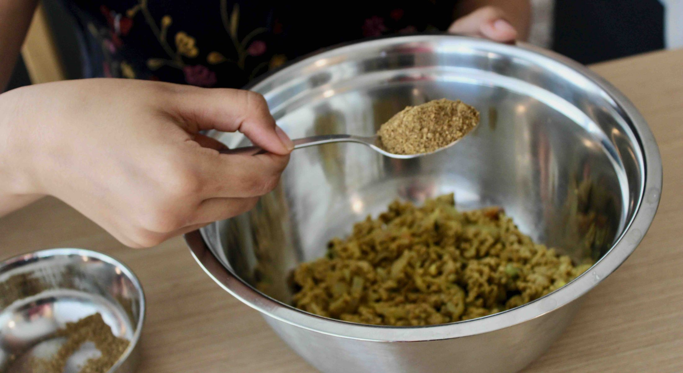 A woman&rsquo;s hand holds a spoonful of brown-coloured spice blend over a metal bowl with food inside it.