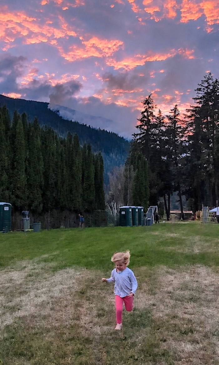 A young child runs on a stretch of green gas at sundown. Behind her, smoke is visible on a mountainside.