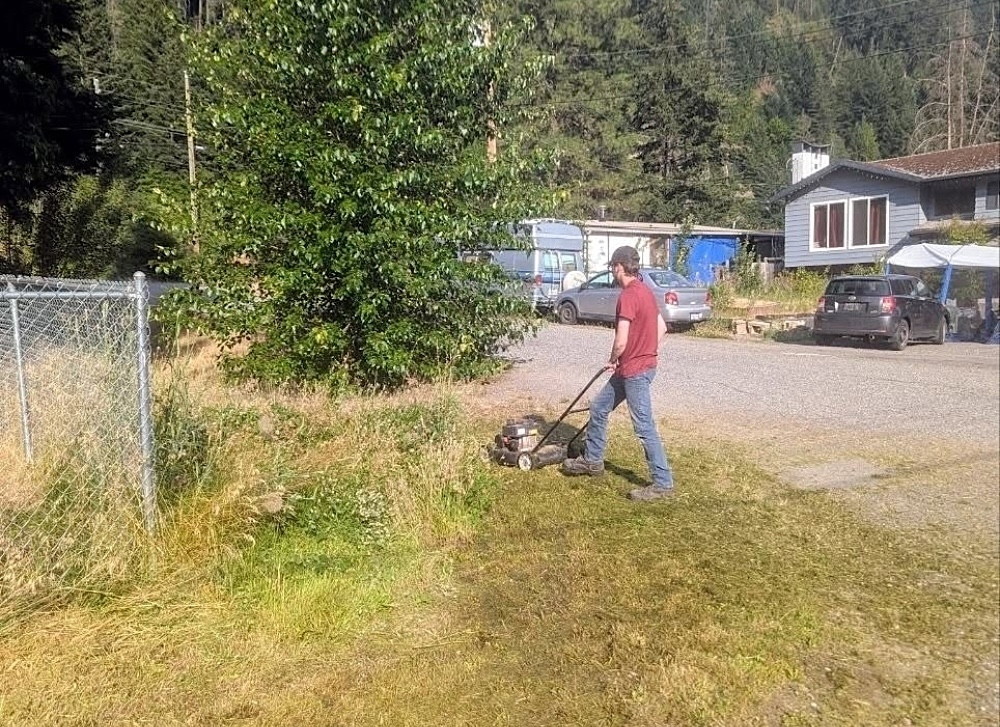 A man mows grass on the side of a gravel road.
