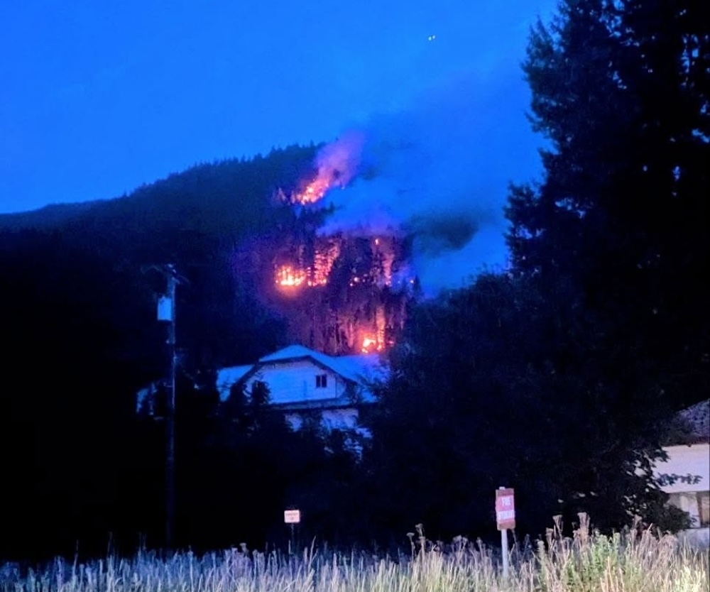 Flames are visible above a white home on a forested mountainside.