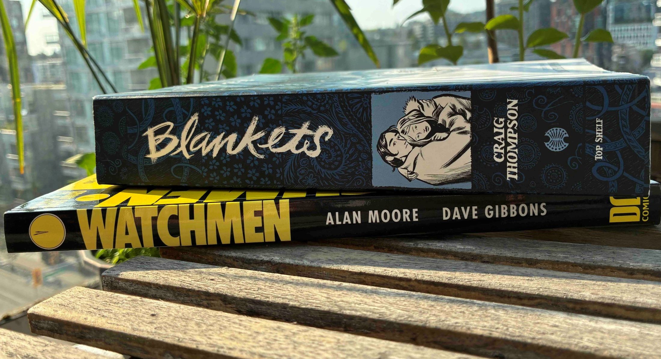 Two graphic novels, &ldquo;Blankets&rdquo; and &ldquo;Watchmen&rdquo;, are stacked together on a small outdoor wooden table. There are green shoots and residential highrises in the background.