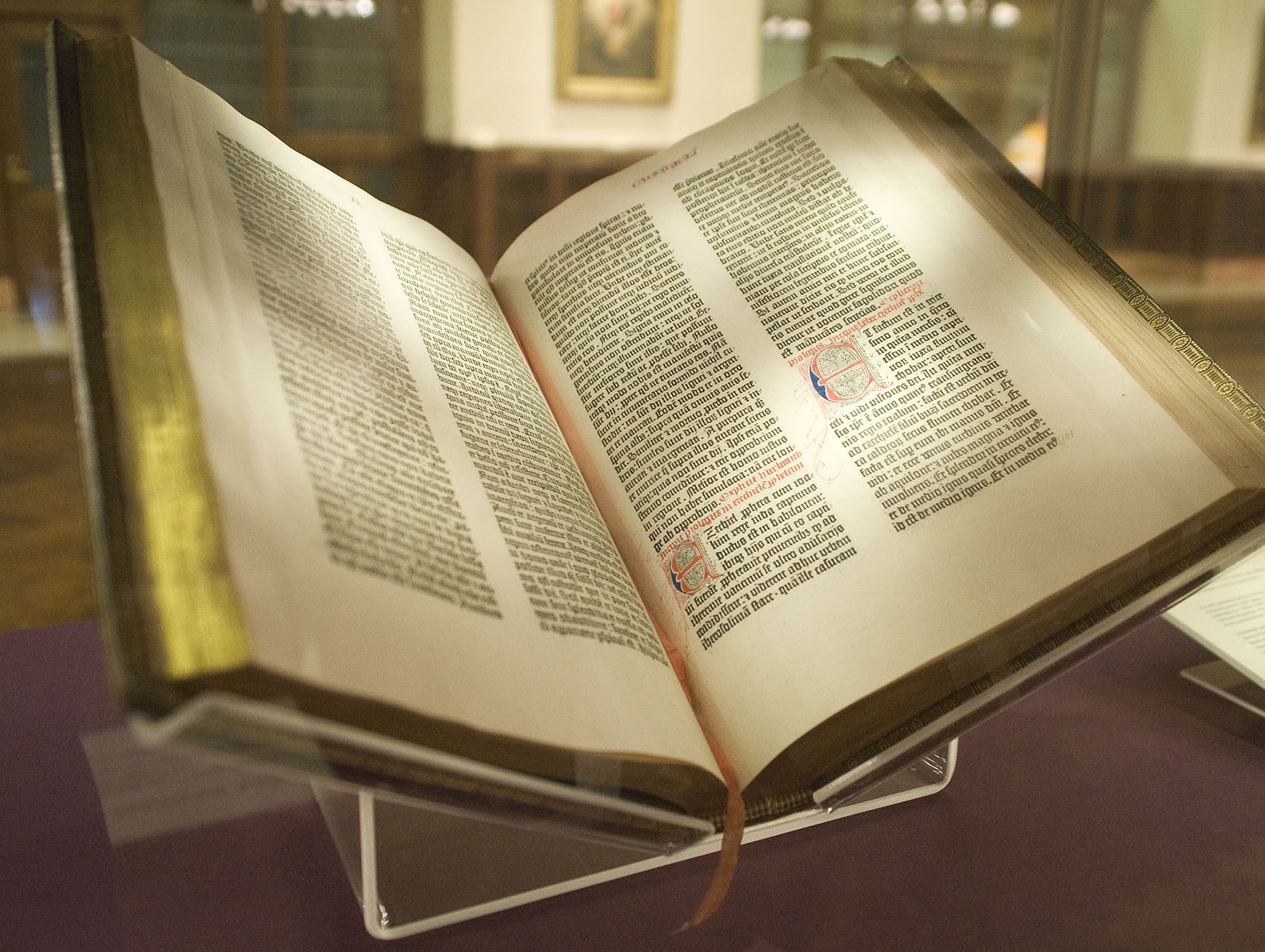 An ancient edition of the Gutenberg Bible sits open on a clear plastic stand. The edges of its pages are gold. 