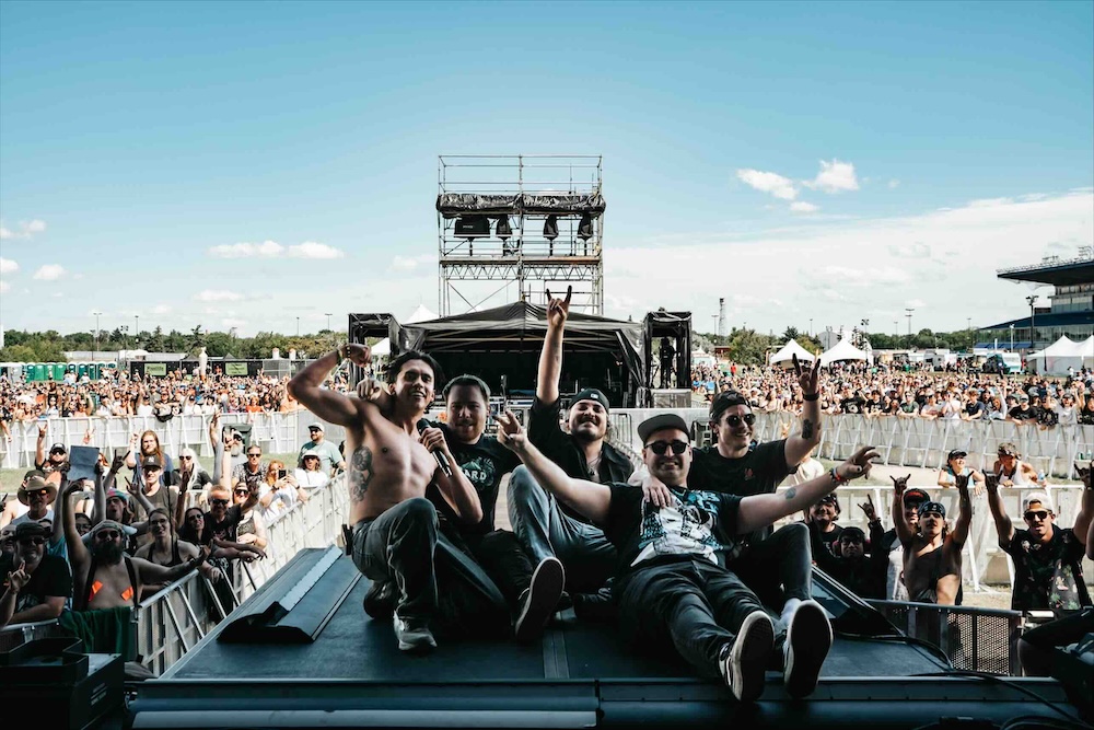 A group of young men poses together on a black, shaded outcropping of an outdoor stage. Crowds of people pose behind them in the sun.