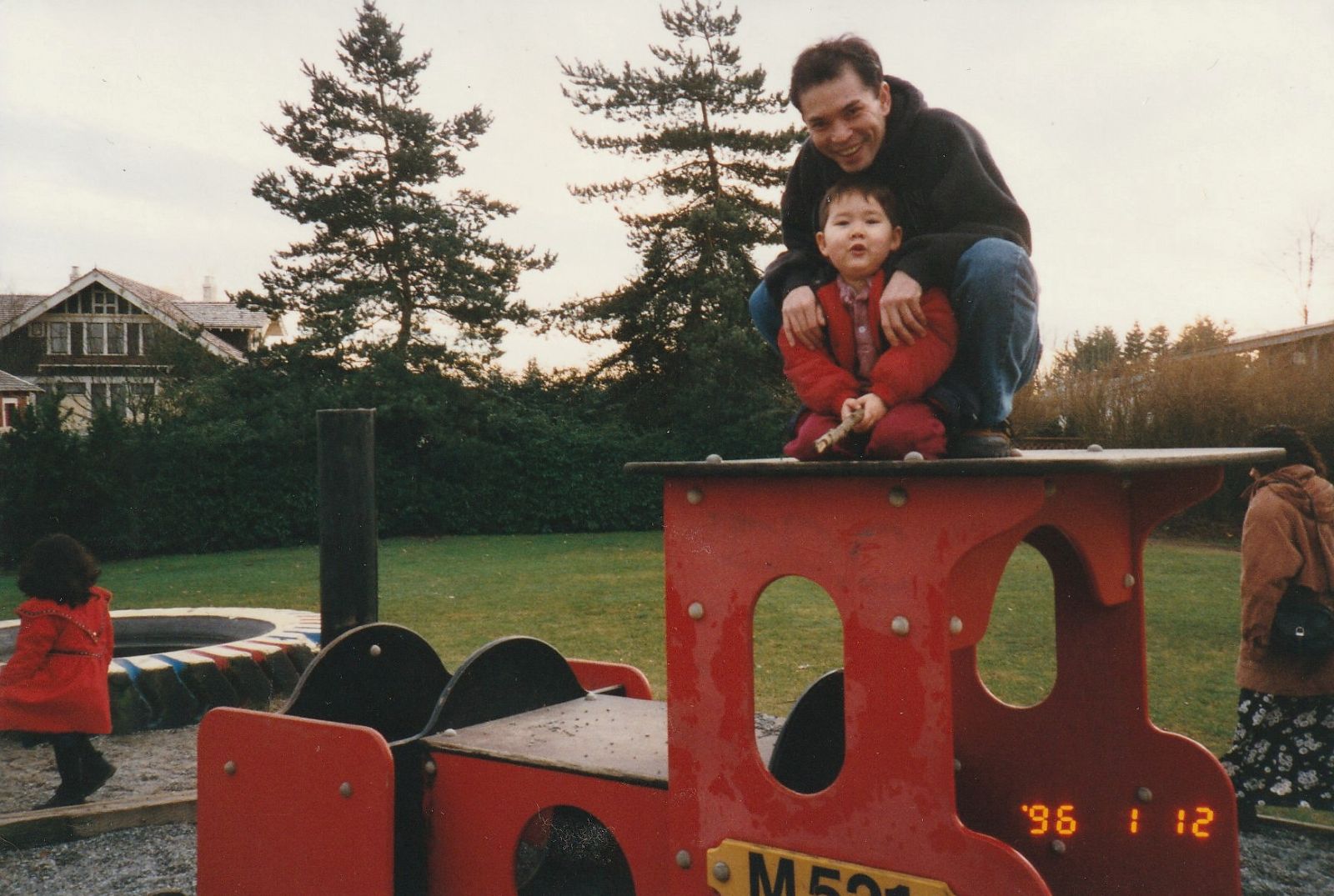 A young boy in a red outfit sits atop a piece of red vehicle-shaped playground equipment in a grassy park with his father, who crouches behind him with his arms around his son&rsquo;s shoulders. The father is wearing a black hoodie and jeans. They are smiling at the camera.