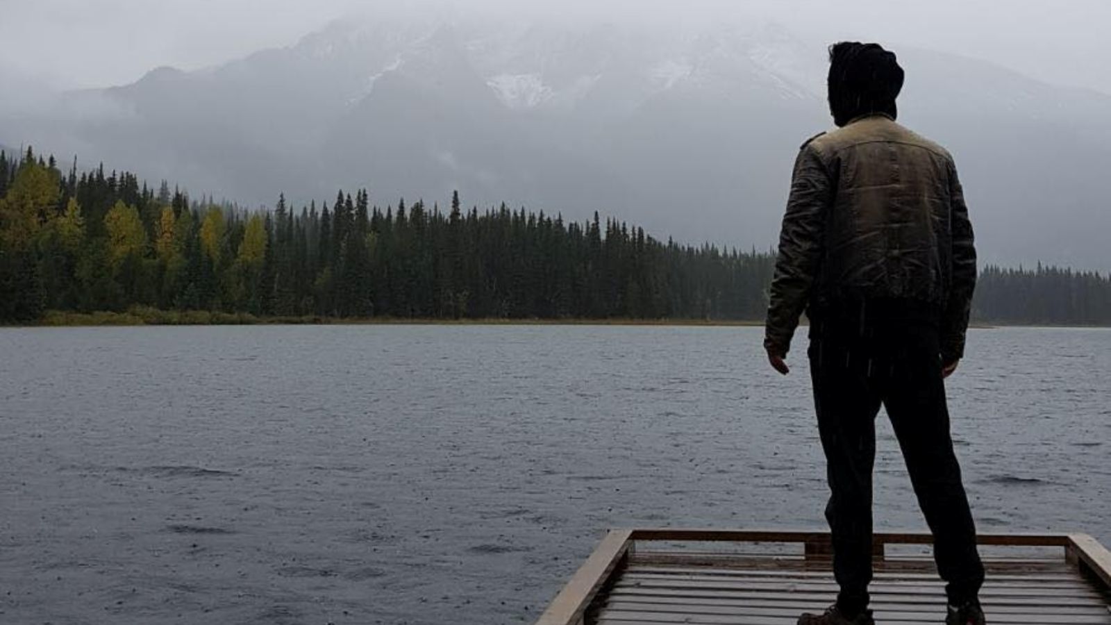 A young man in a dark hoodie and jacket stands with his back to the camera on the dock of a body of water on a grey day. In the background is a stand of coniferous trees.