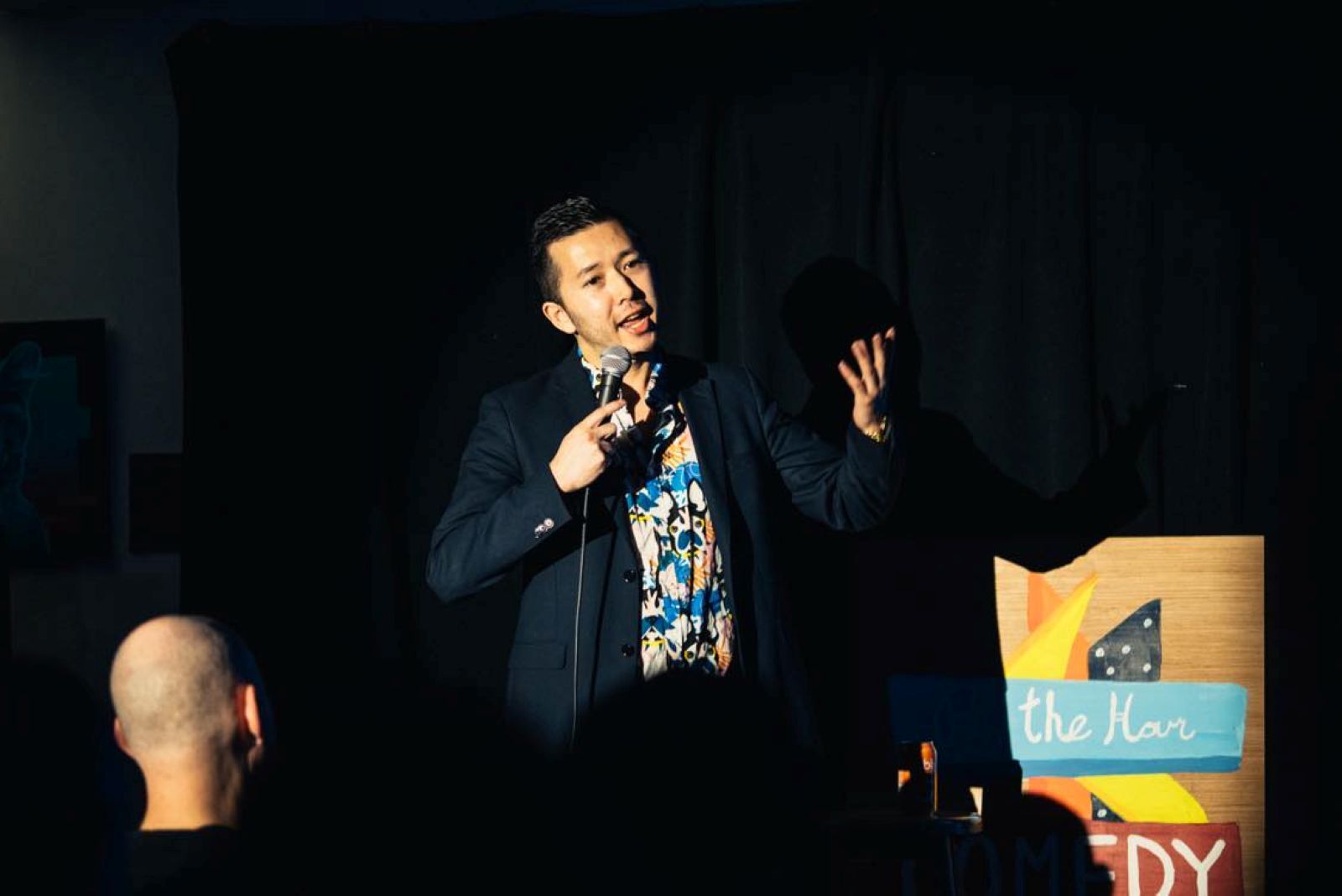 Trevor Jang stands onstage before a crowd. He is speaking into a microphone and gesturing with his left hand. He has short dark hair and wears a navy blazer over a colourful patterned shirt.