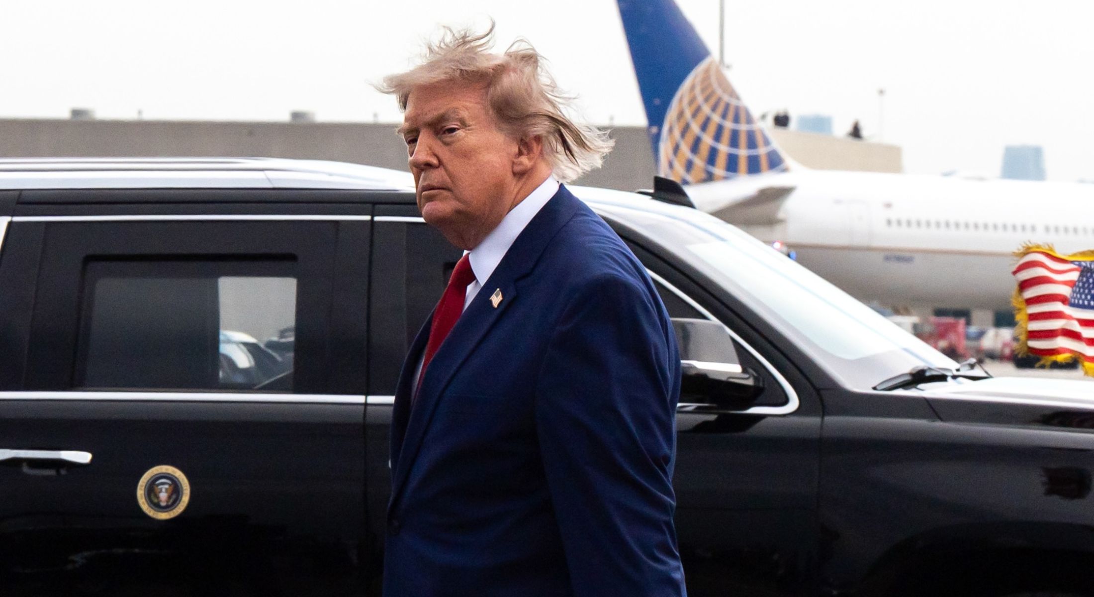 President Donald Trump walks on the tarmac of an airport in front of a black presidential limousine. He is wearing a navy suit with a red tie. His hair blows in the wind.