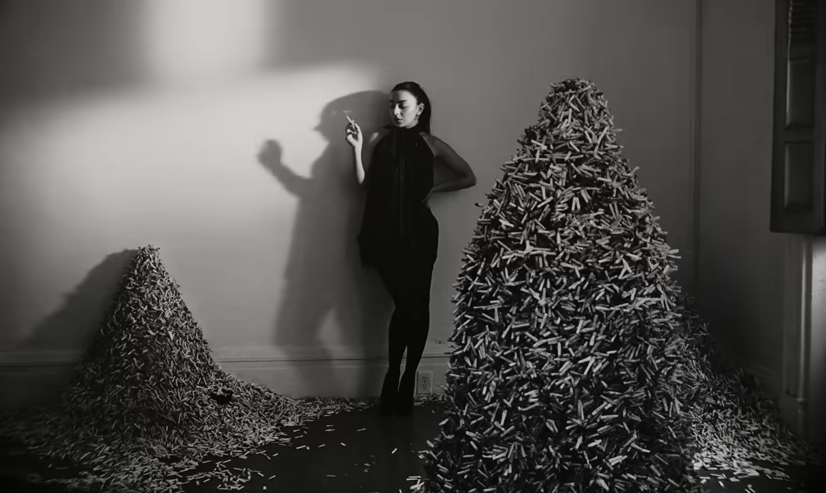 A black-and-white photograph depicts a young woman in a ponytail smoking a cigarette in a room stacked with tall piles of cigarettes.